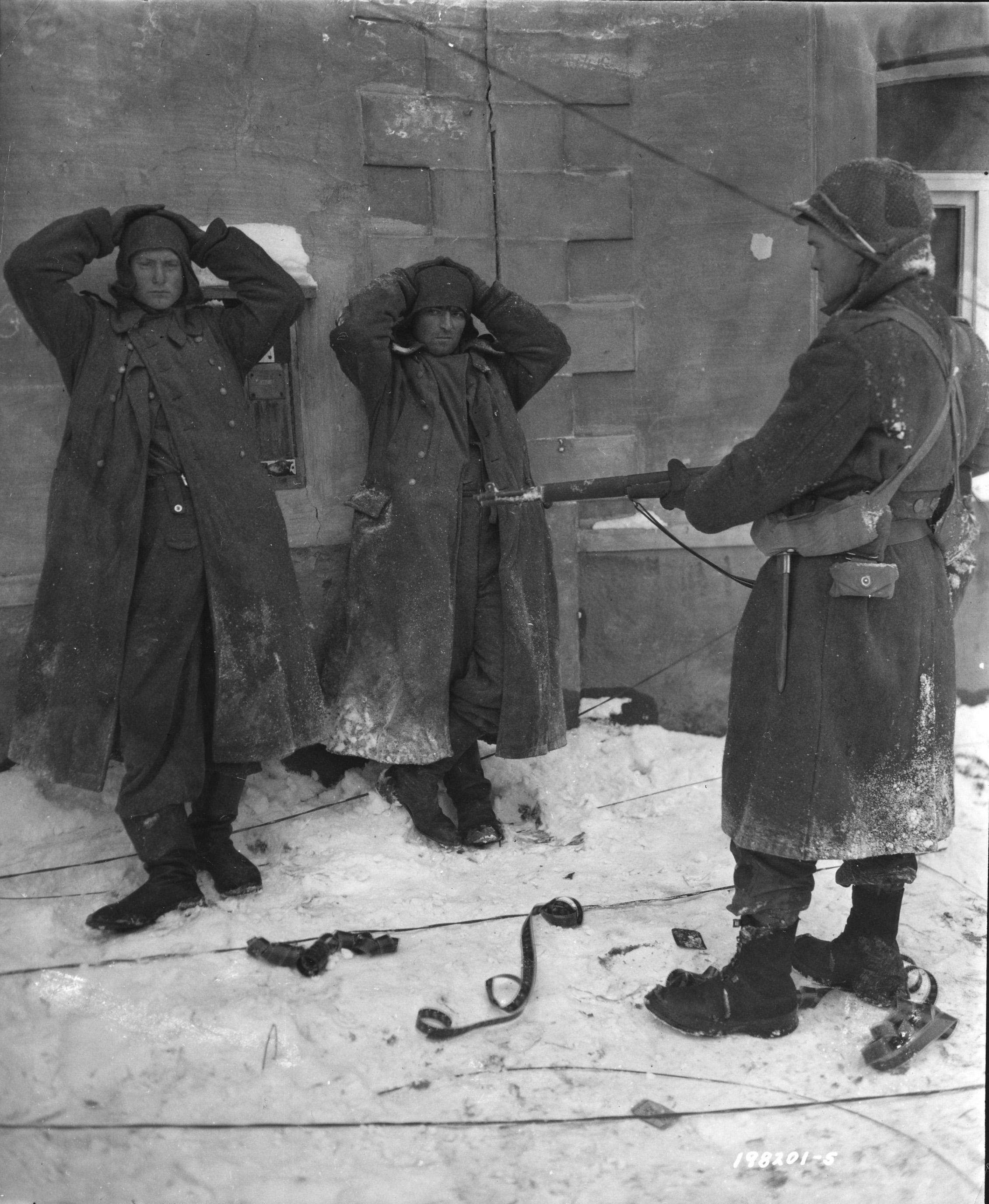 Two German SS soldiers are standing against a wall, while a US soldier guards them.