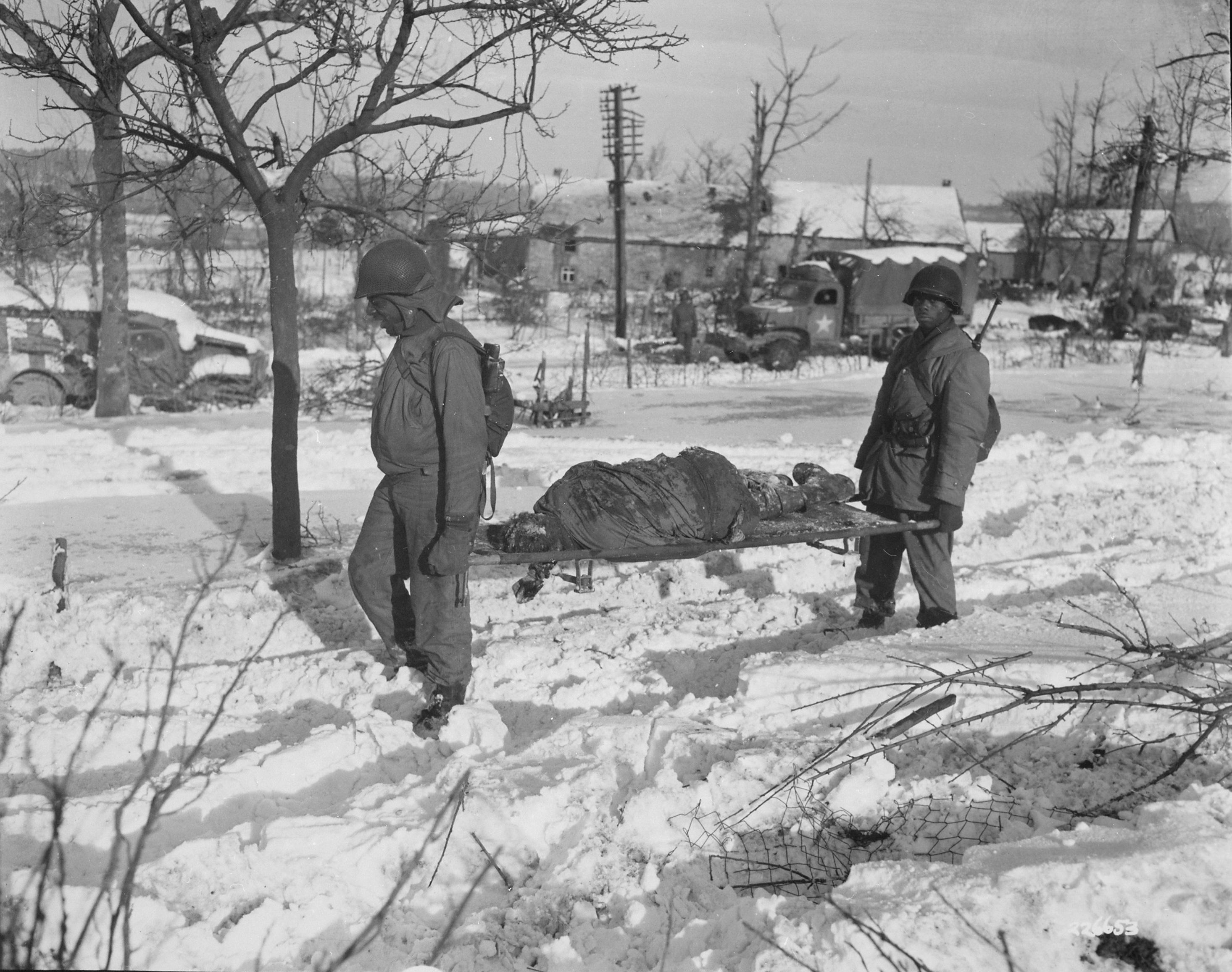 A photograph of two Black American soldiers carrying away a victim of the Malmedy Massacre on a stretcher.