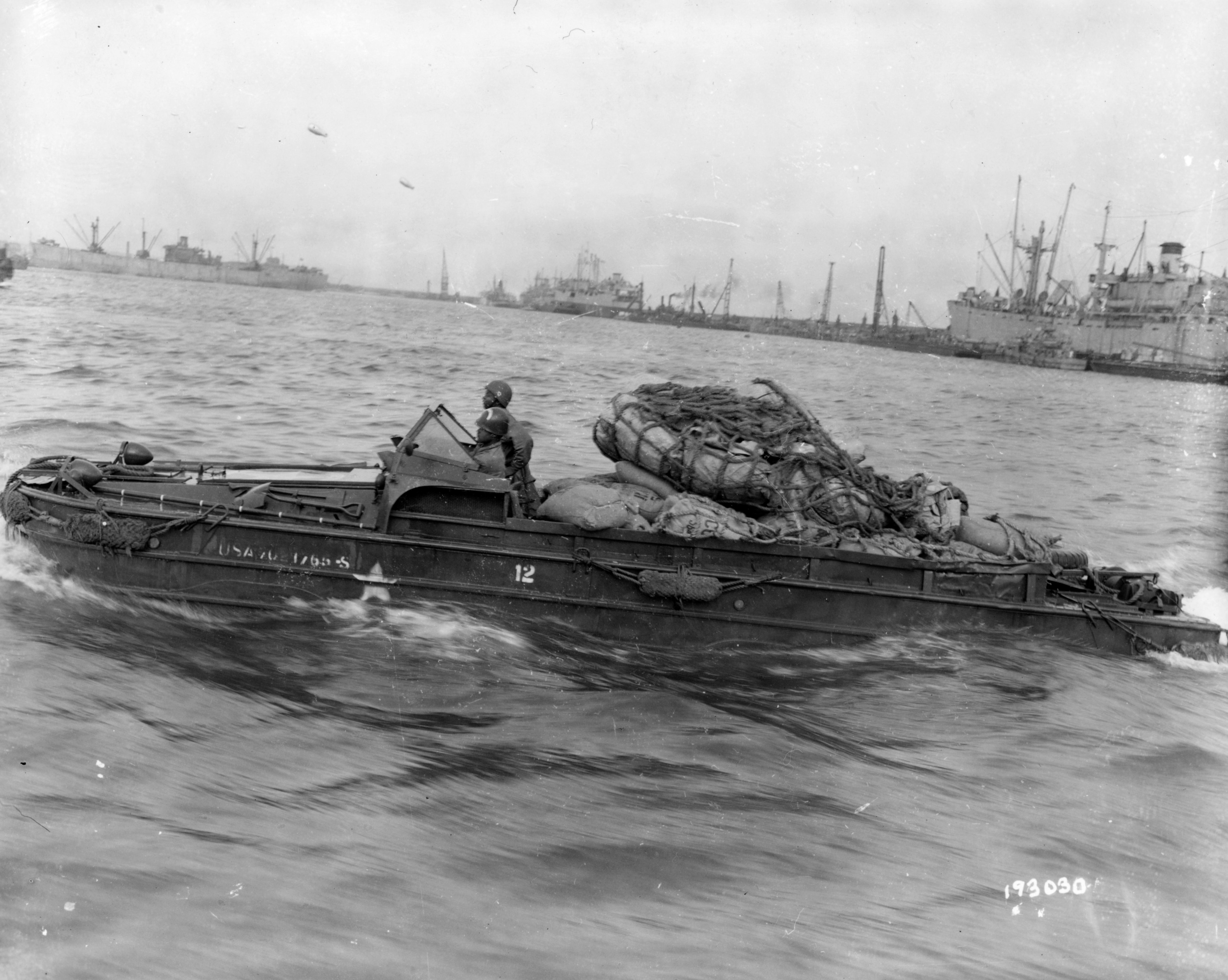 Historical image showing A DUKW operated by men of a port battalion carries cargo from Cherbourg harbor to mainland France. of Black American soldiers during WWII