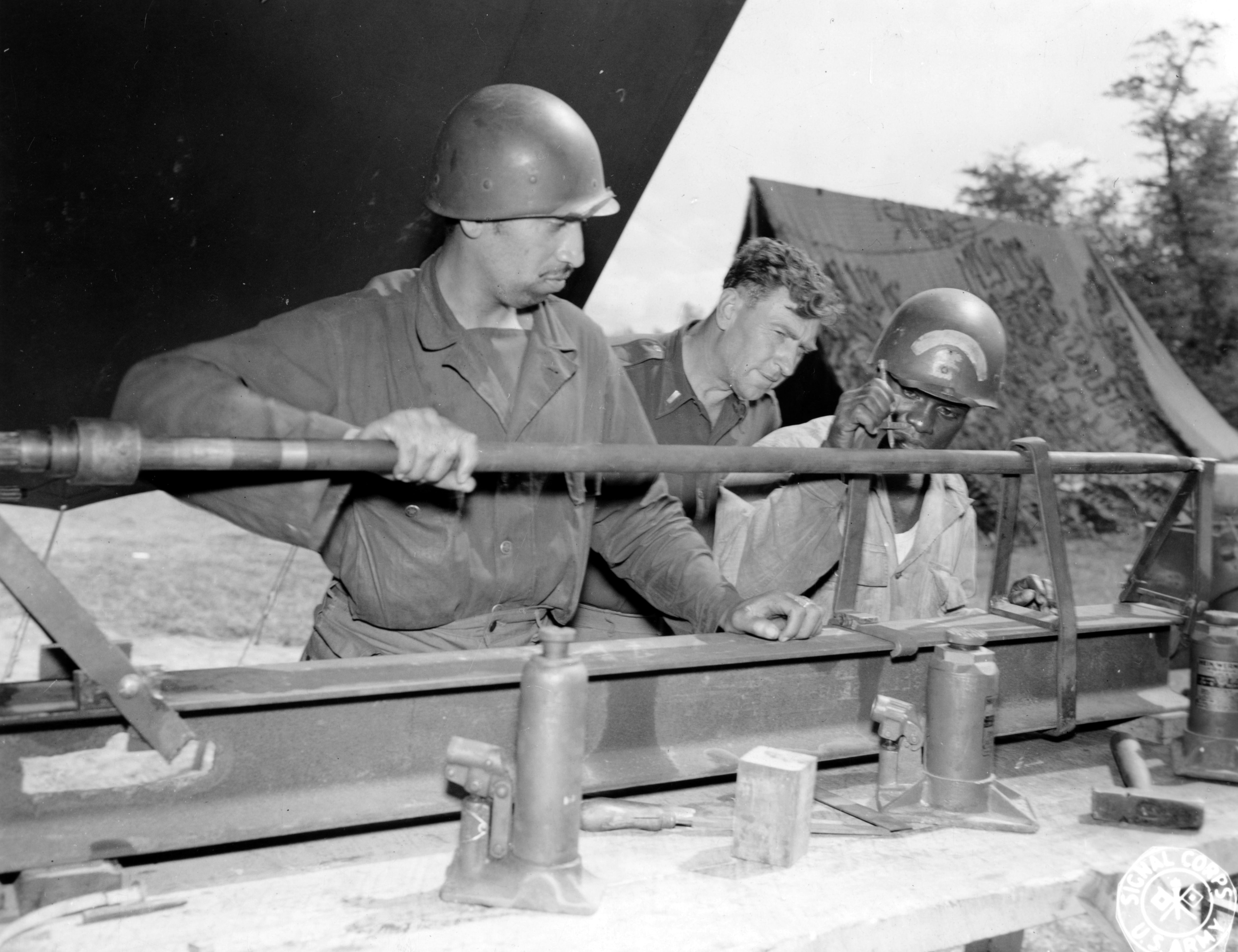 Historical image showing Sgt. Albert R. Jenkins, 1st Lt. Loren J. Pryor, and Sgt. Junius Meade straighten a propeller shaft of a DUKW. of Black American soldiers during WWII