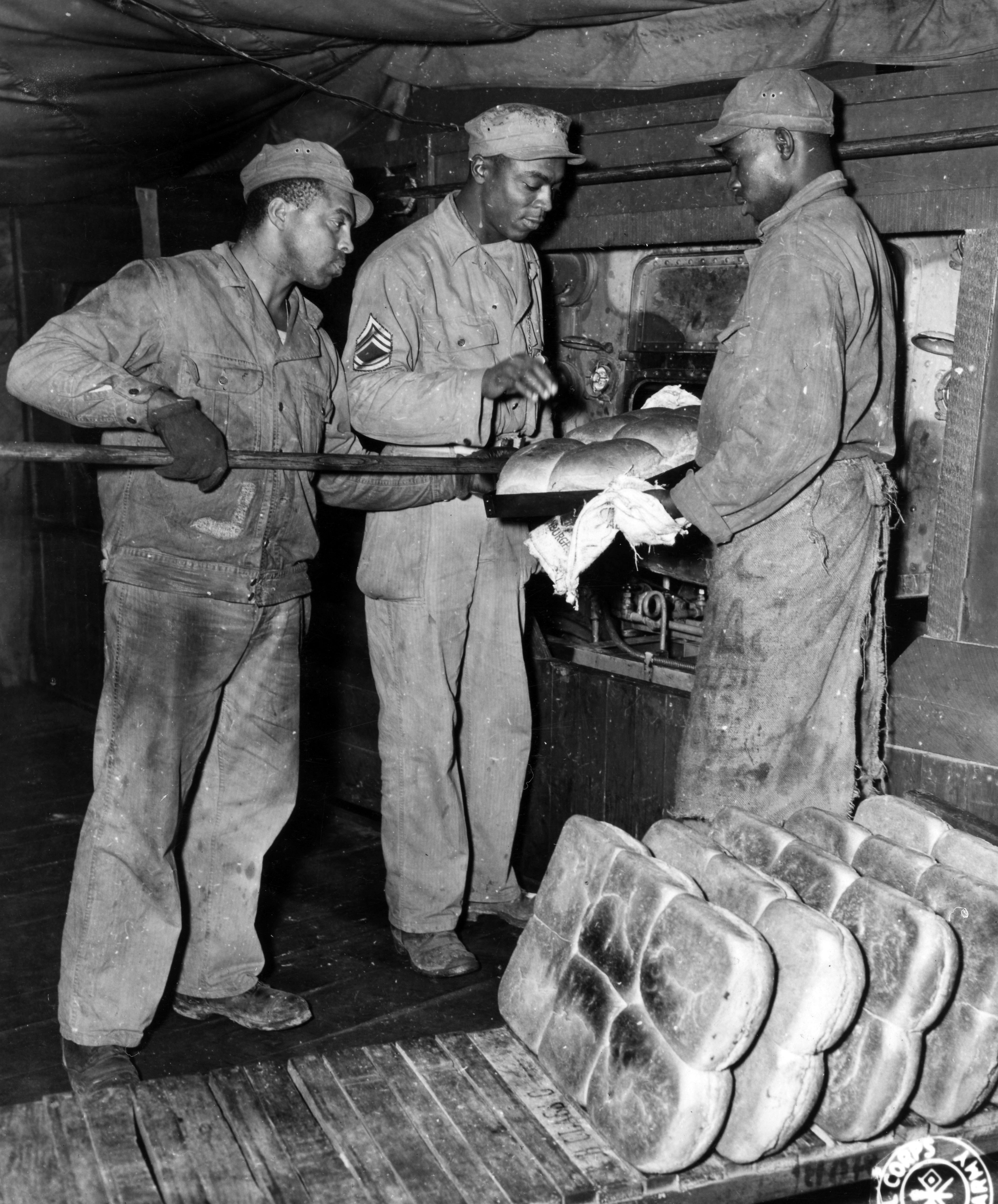 Historical image showing Three bakers at a depot, somewhere in England, test a fresh pan of loaves as they come out of the oven. of Black American soldiers during WWII